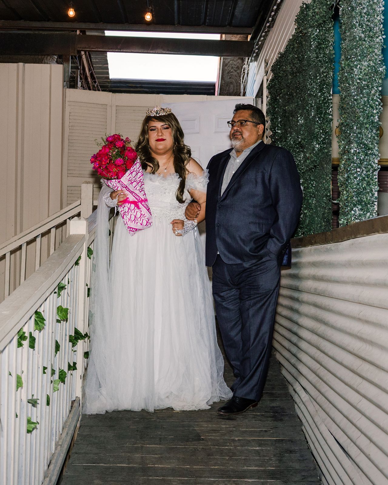 Bride in white gown with pink bouquet and tiara stands with man in navy suit on decorated staircase.