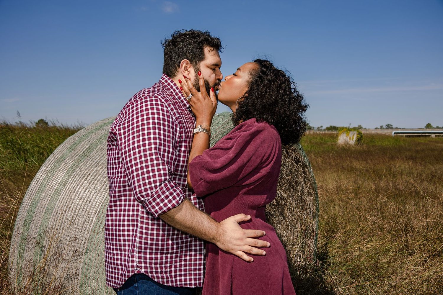 A couple shares a romantic kiss in front of a round hay bale in a golden field on a sunny day.