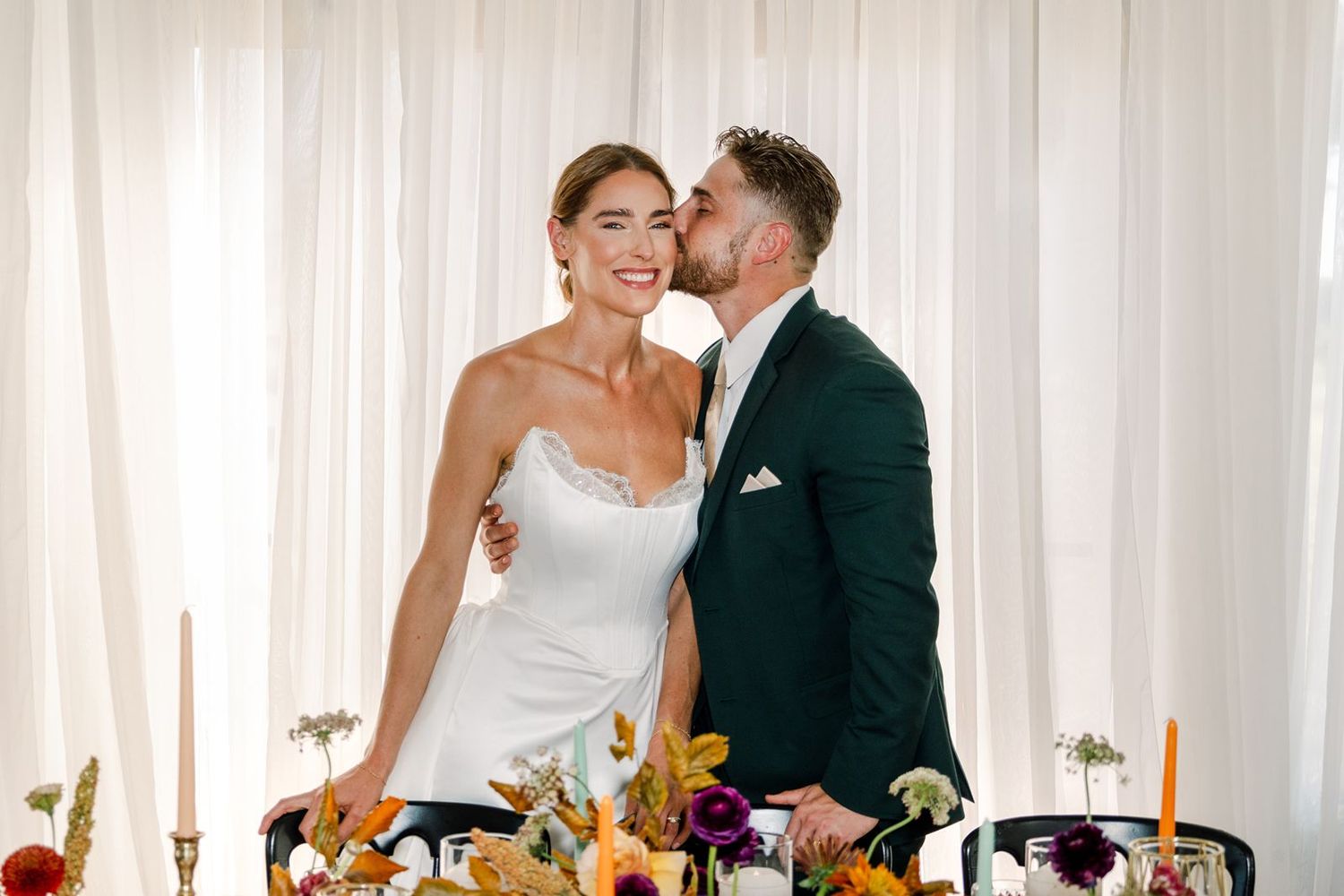 A couple in wedding attire shares a joyful moment at their reception table decorated with elegant candles and florals.