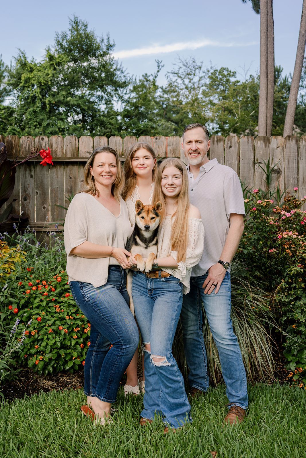 A family poses together with their corgi dog in a backyard garden setting with blooming flowers.