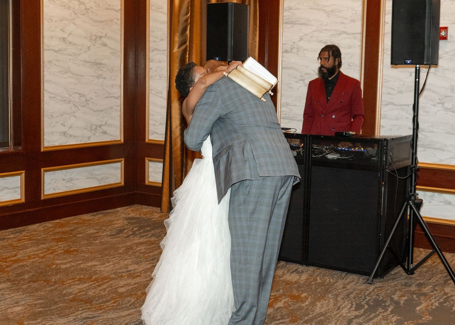 Bride and groom share a tender embrace on the dance floor with DJ performing in background.