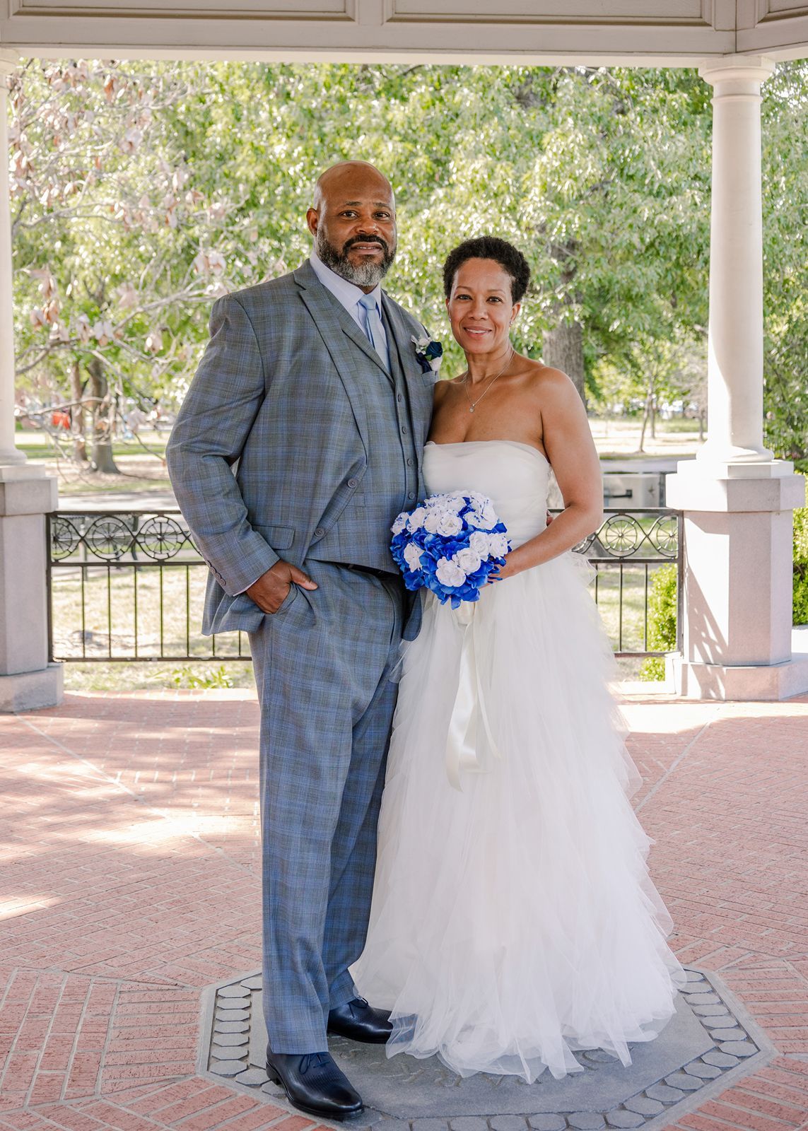 Wedding couple standing under gazebo, groom in gray plaid three-piece suit, bride in strapless white gown with blue bouquet.