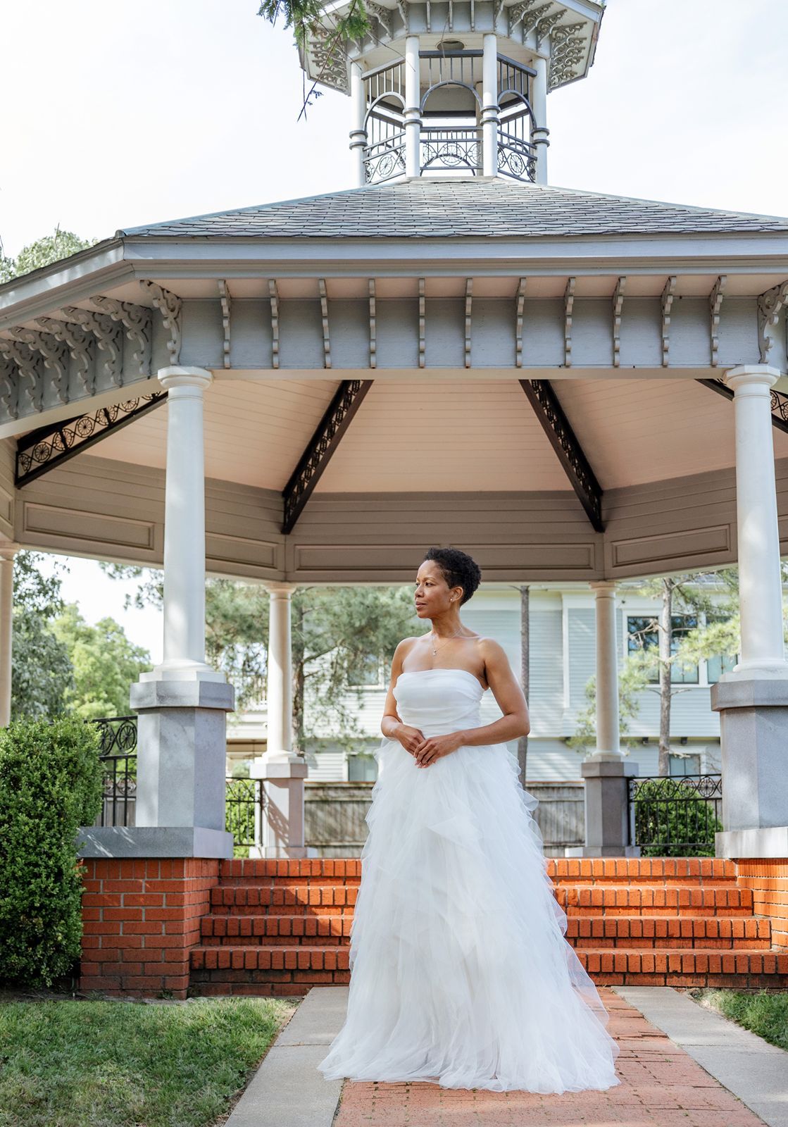 Bride in strapless white layered gown standing before a classic gazebo in an outdoor wedding setting.