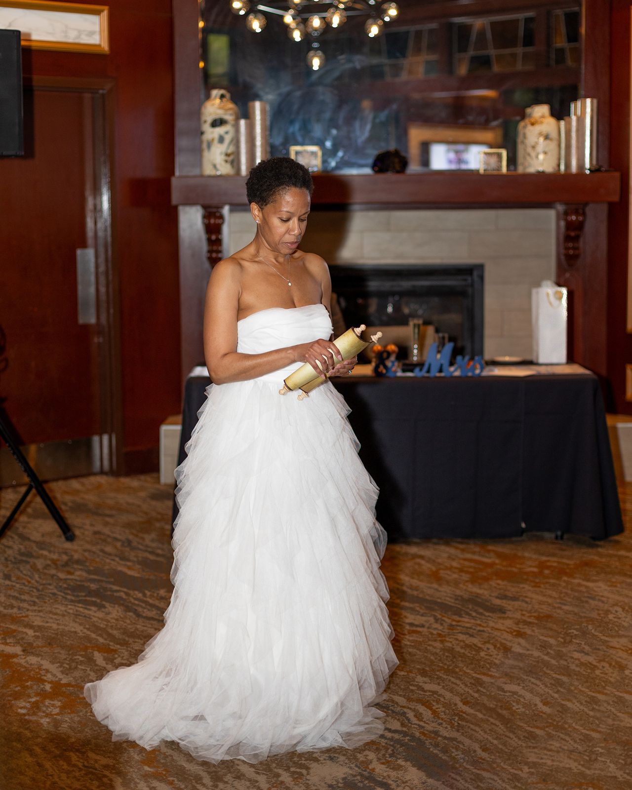 Bride in strapless white ruffled wedding gown holding gold item near fireplace.