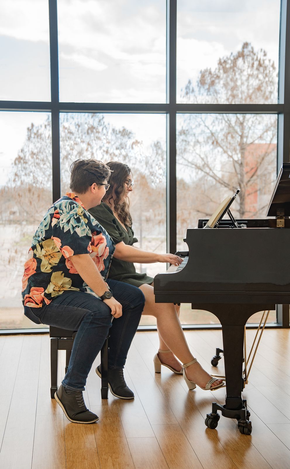 Two people sit together at a grand piano near large windows with a view of bare trees outside.