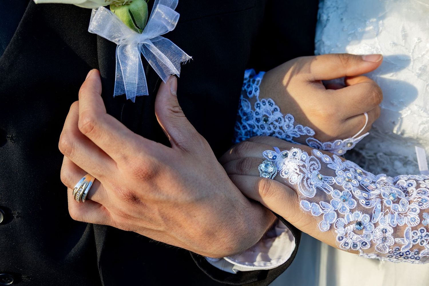 Close-up of bride and groom hands with elegant lace glove, pearl ring, and boutonniere at wedding ceremony.
