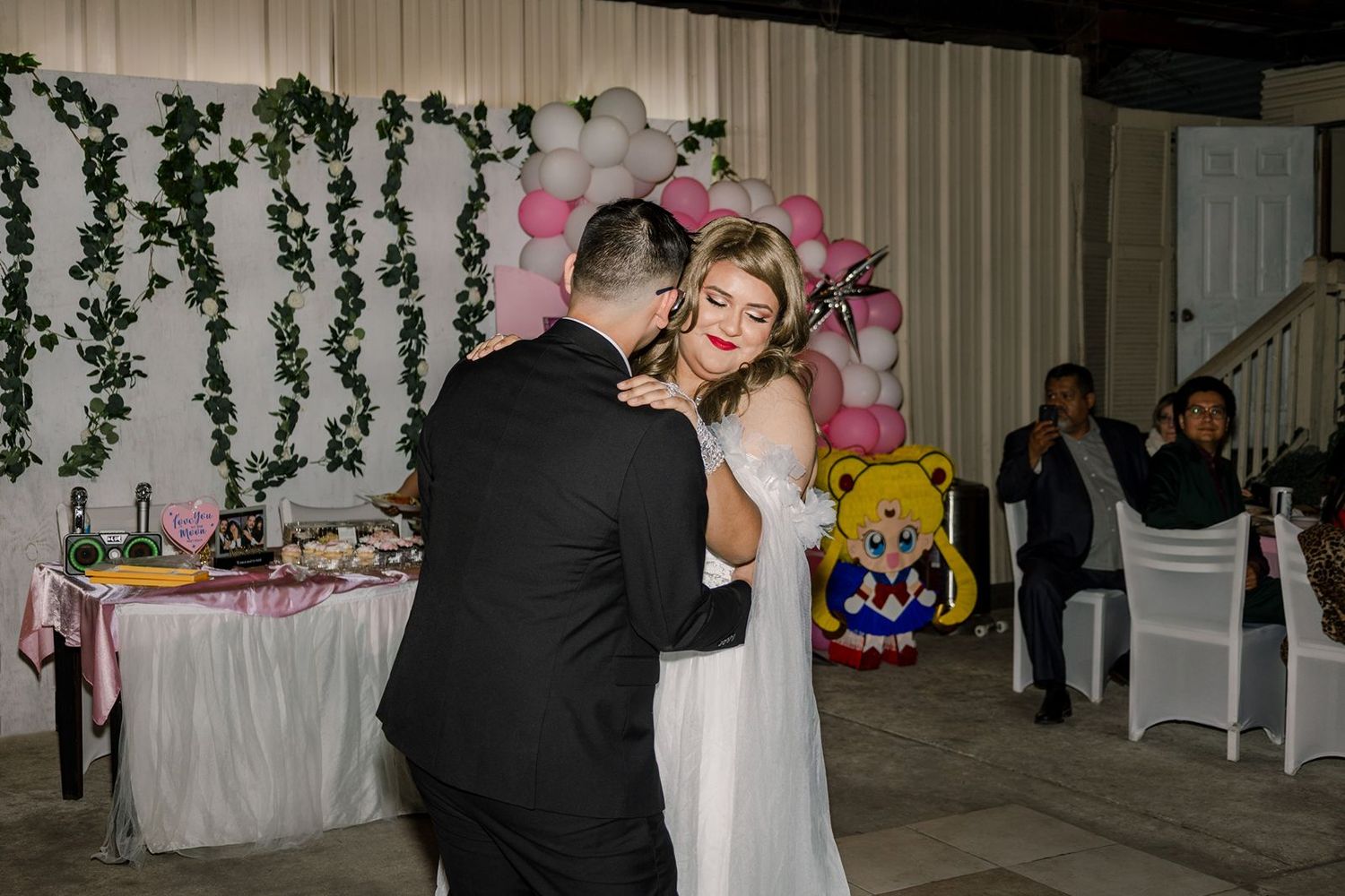 Couple shares first dance at wedding reception with pink balloon arch and Sailor Moon decoration in background.