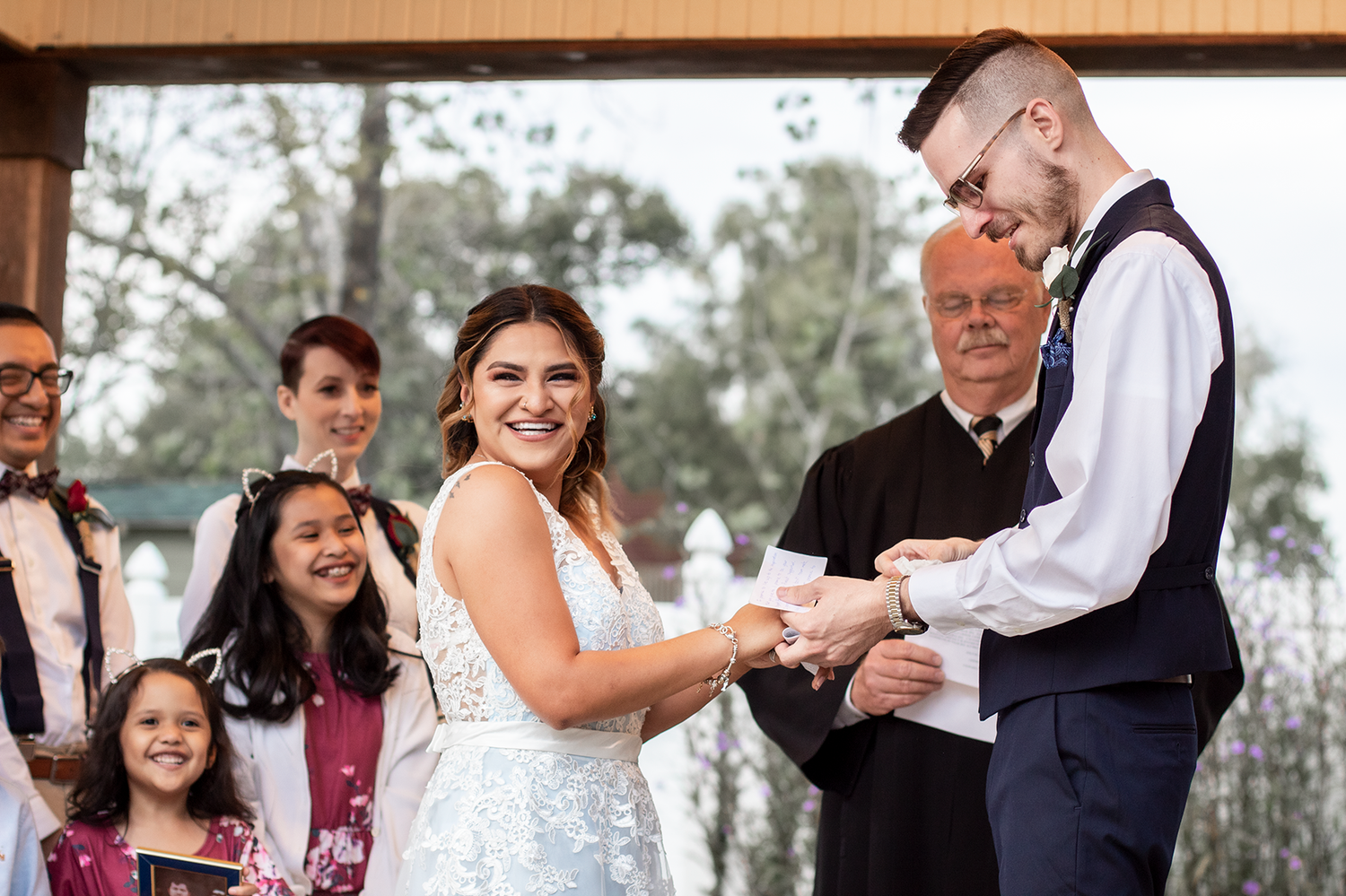 Bride and groom exchanging rings during outdoor wedding ceremony with officiant and guests smiling in background.