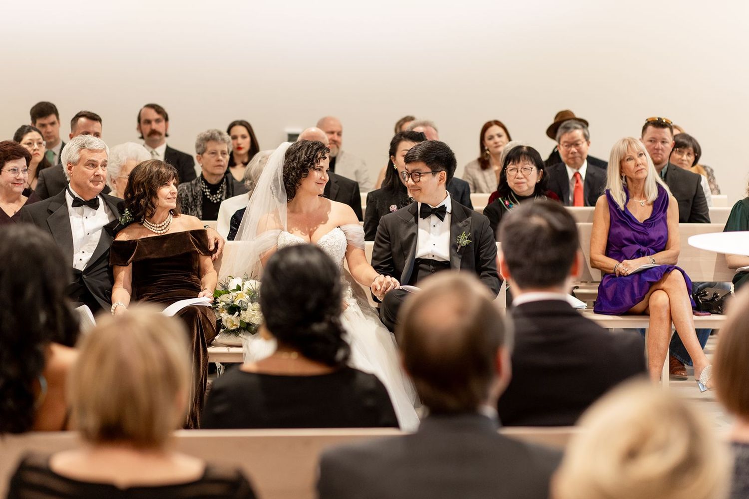 Wedding guests seated in rows watch attentively during an indoor ceremony celebration.
