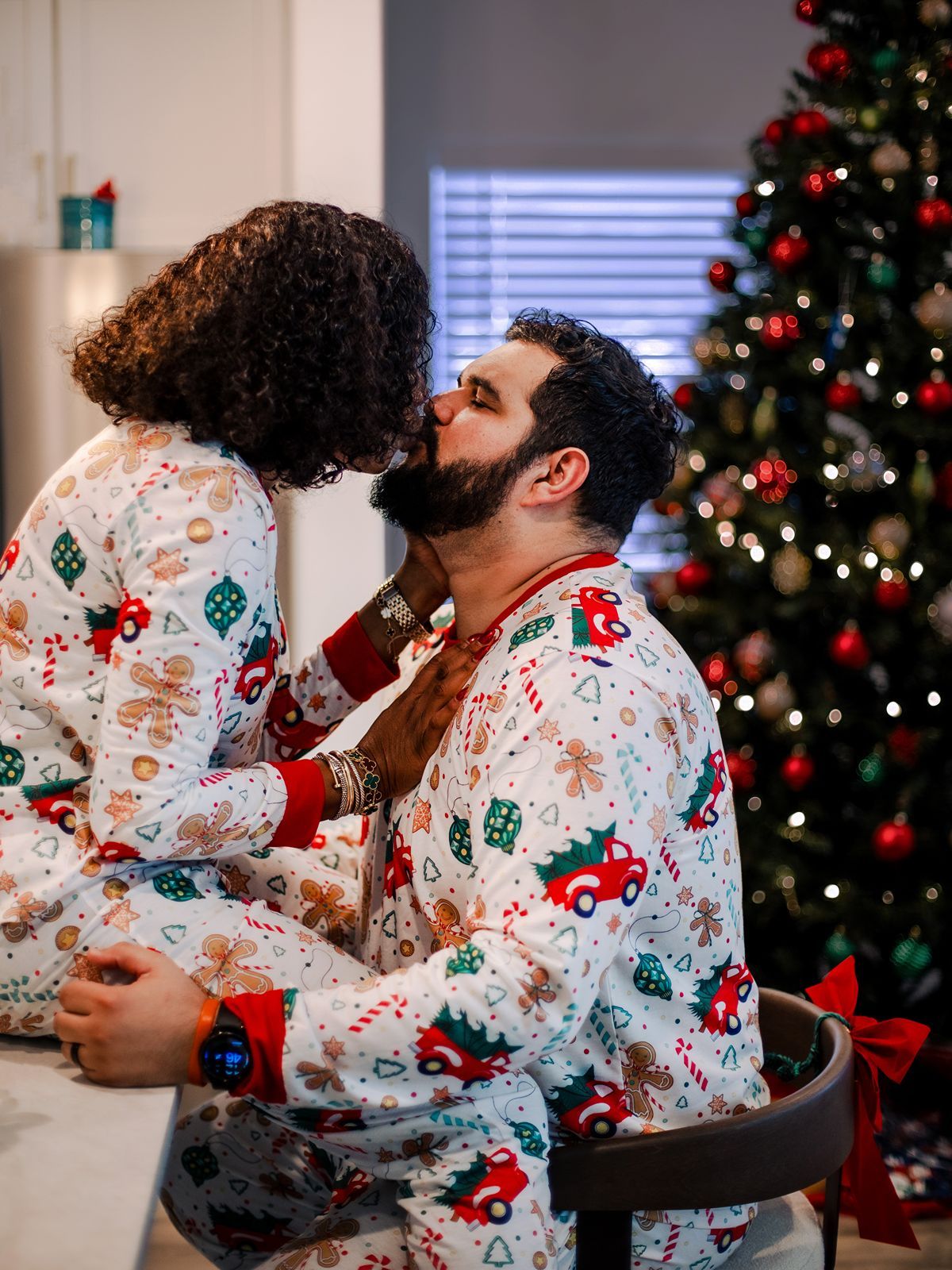A couple wearing matching Christmas pajamas share an intimate moment in front of a decorated tree.