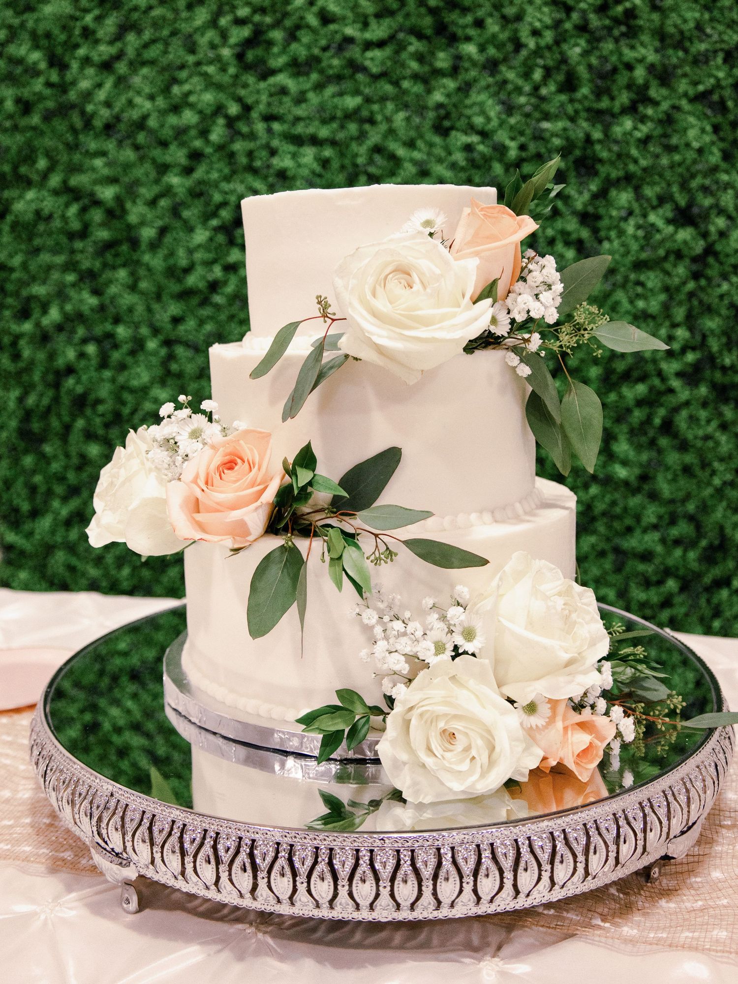 Elegant white wedding cake decorated with fresh roses and eucalyptus on silver cake stand.
