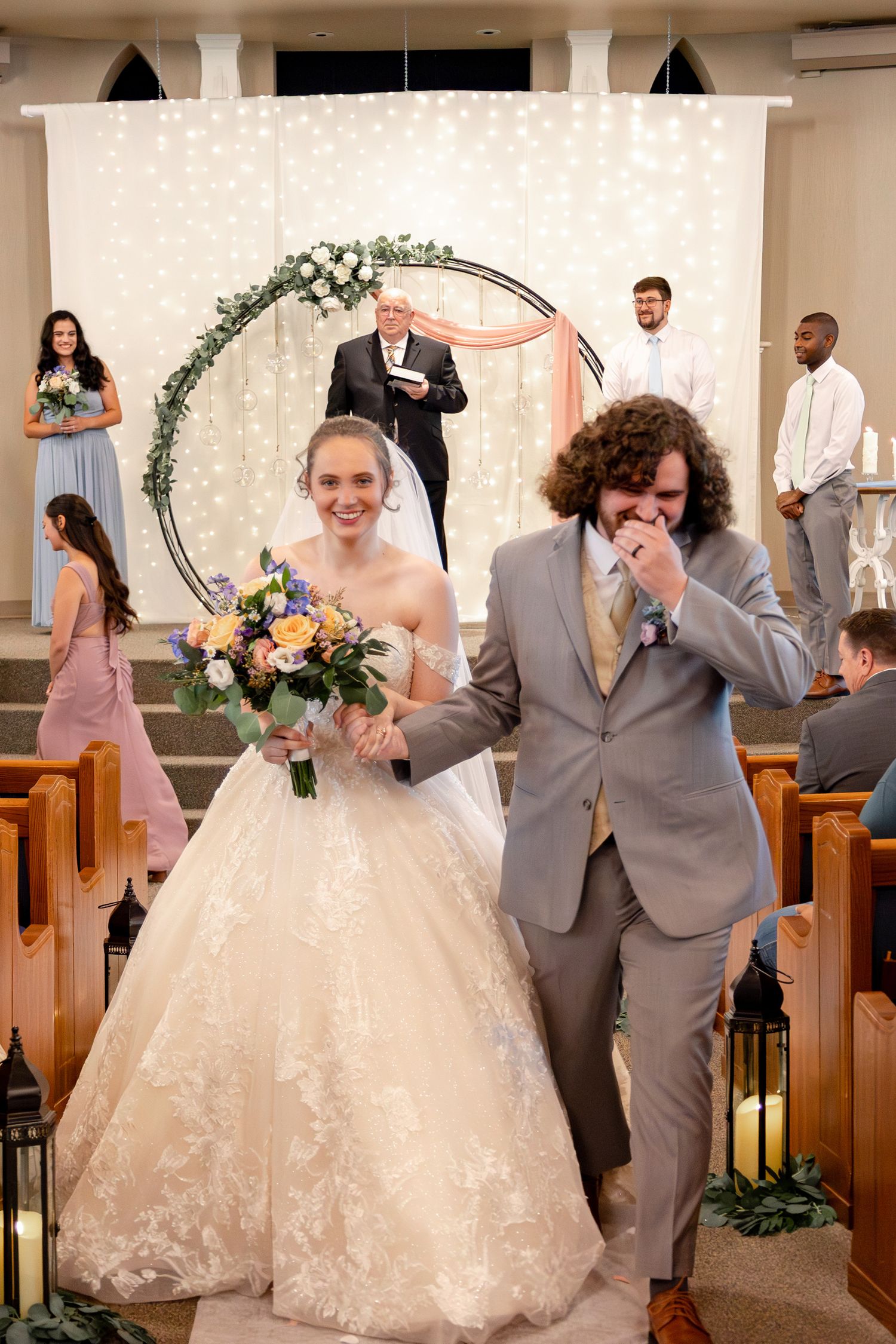 Wedding party celebrating at the altar with decorative arch and white twinkle lights in background.