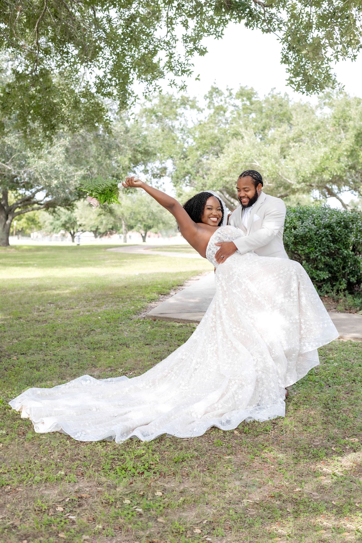 A bride in a flowing white gown twirls joyfully on grassy grounds beneath oak trees.
