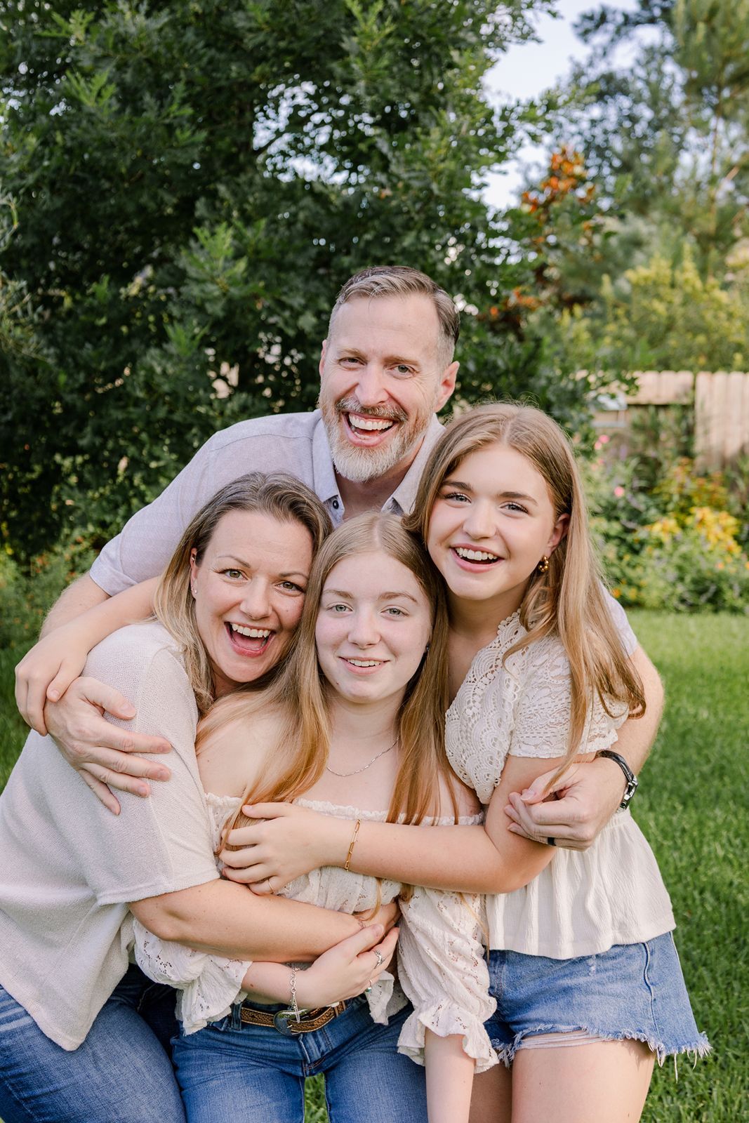 A joyful group embraces and laughs together while posing on grass in a backyard setting.