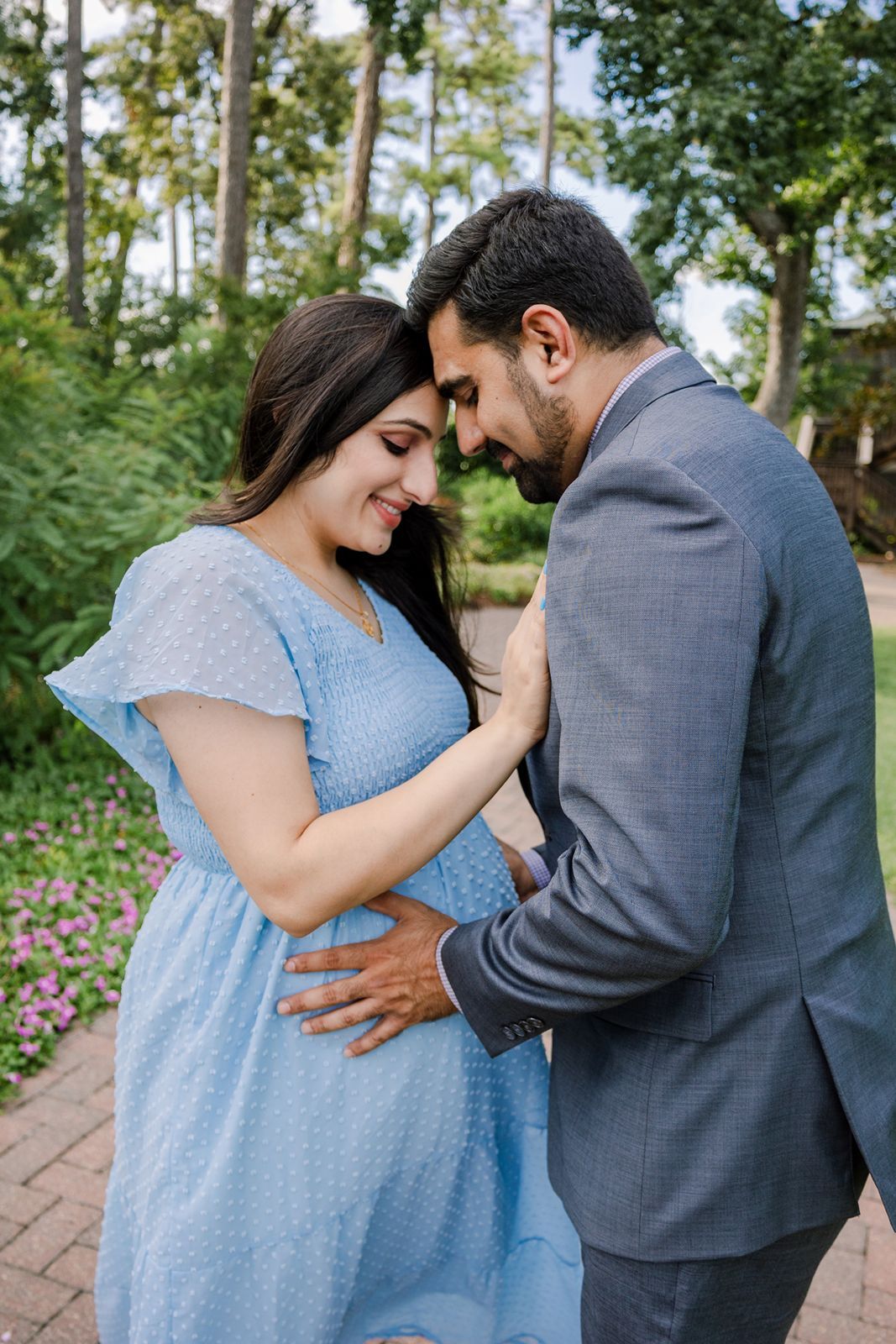 A couple in a garden setting share an intimate moment, with her in a blue dress and him in a grey suit.