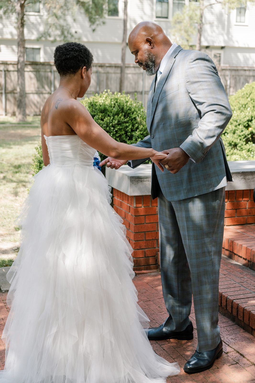 Bride and groom exchanging rings outdoors during wedding ceremony, bride in white tulle gown, groom in gray plaid suit.