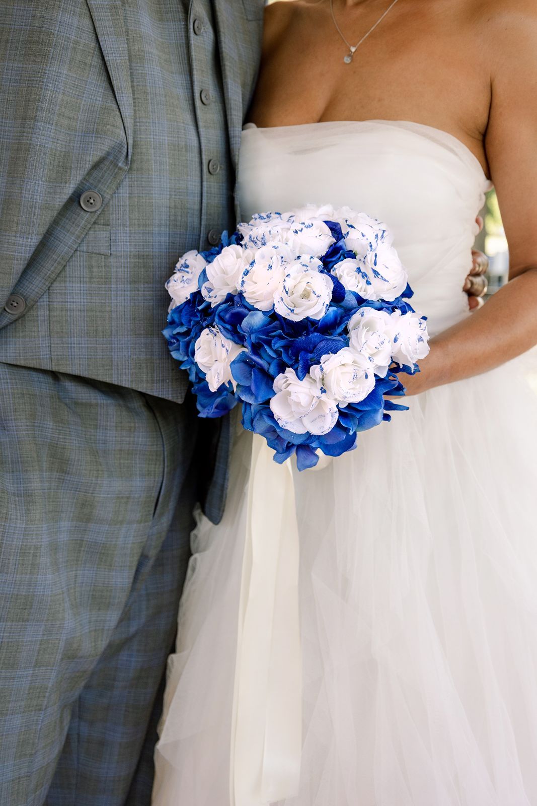 Bride in white strapless gown holds blue hydrangea and white rose wedding bouquet beside groom.