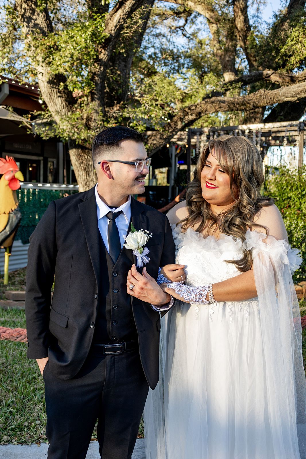 Groom in black suit with boutonniere gazes at bride in white off-shoulder gown at outdoor wedding.