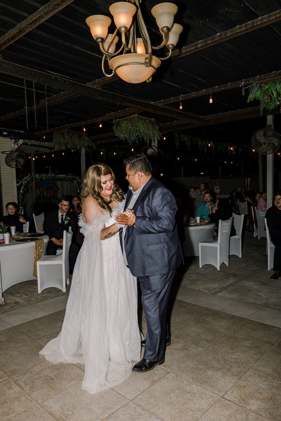 Bride and groom share first dance at outdoor wedding reception with chandelier and string lights.