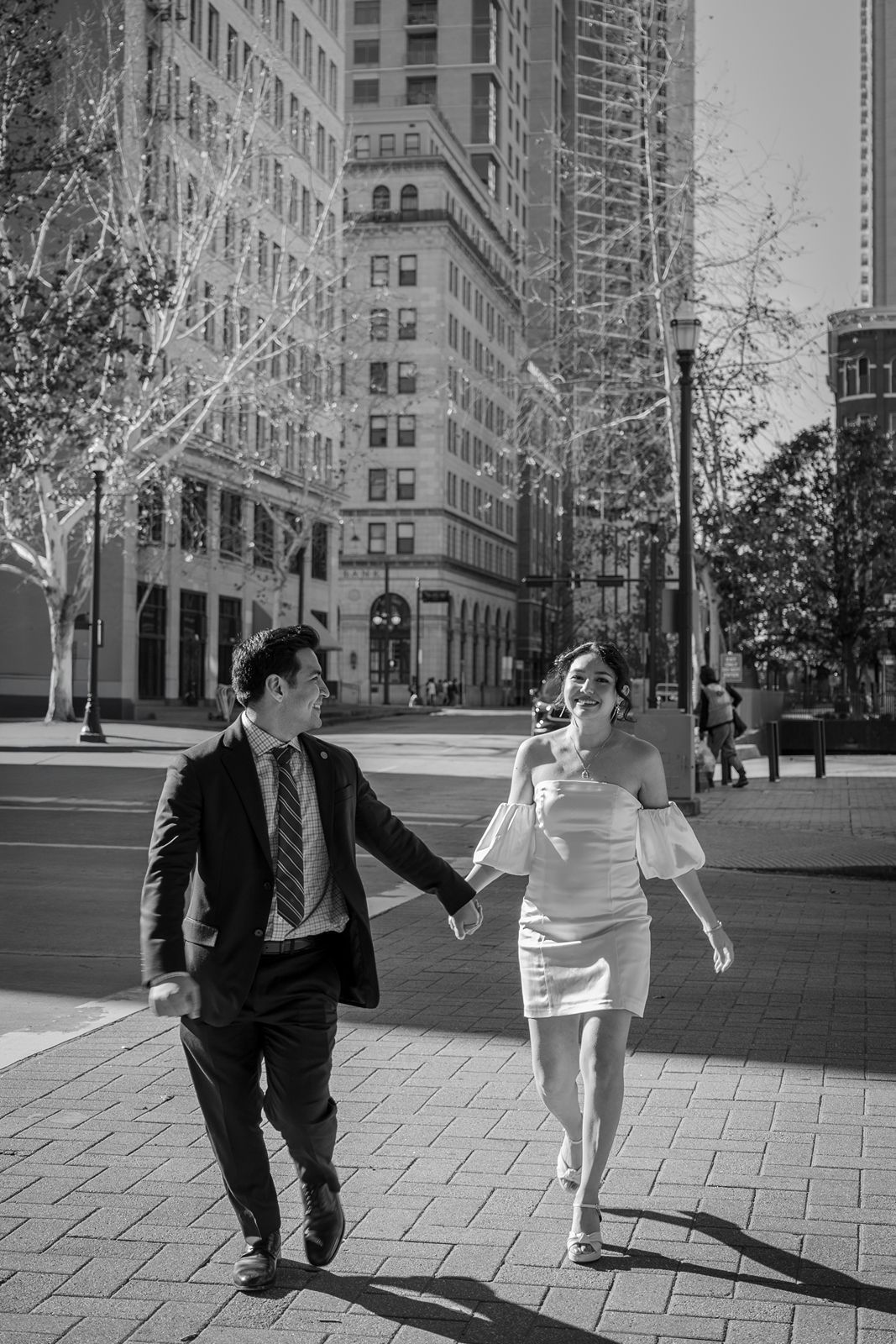 A couple joyfully runs together on a brick sidewalk in a downtown city setting with tall buildings in black and white.