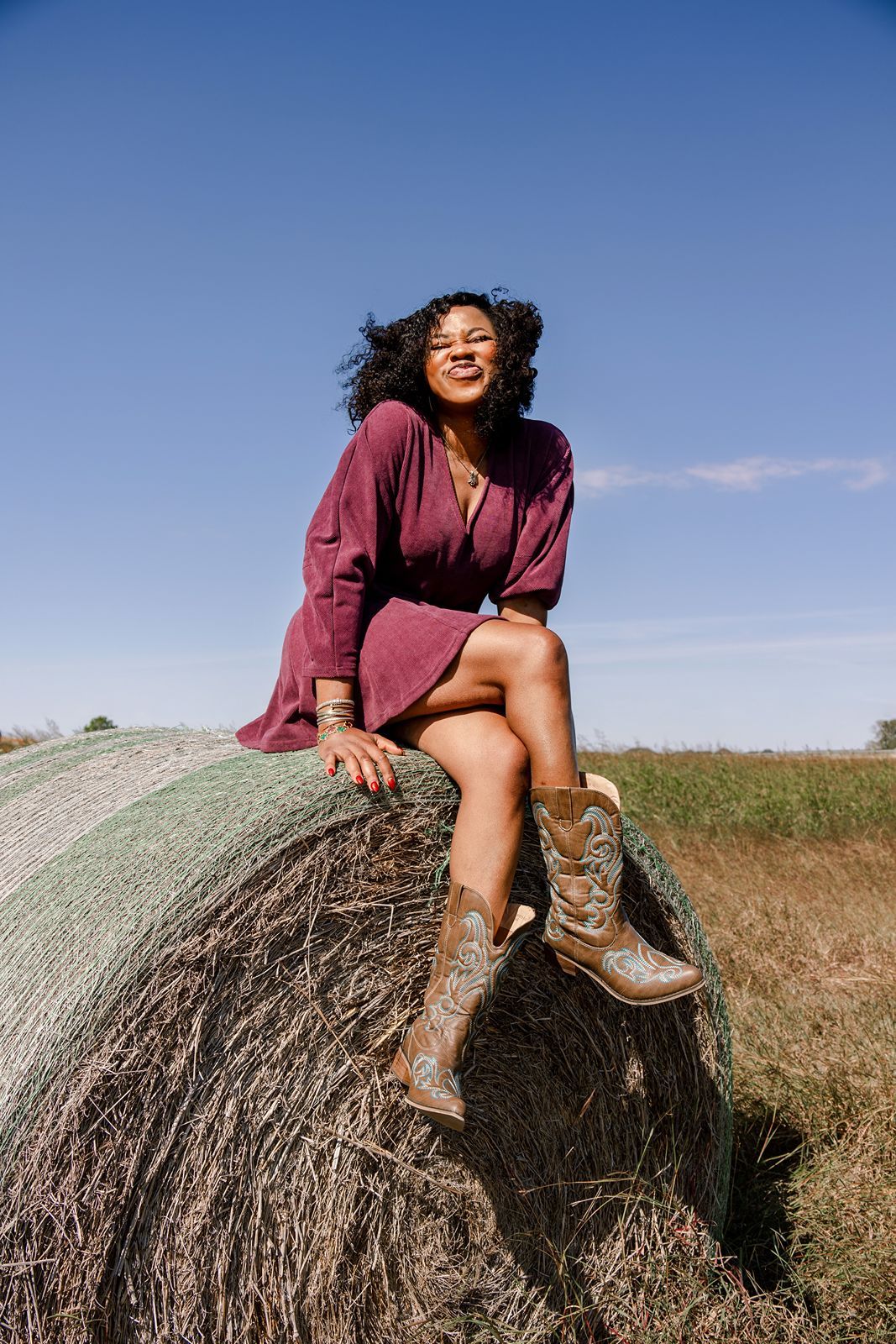 Someone in a burgundy sweater sits casually atop a large round hay bale in a sunny field against blue sky.
