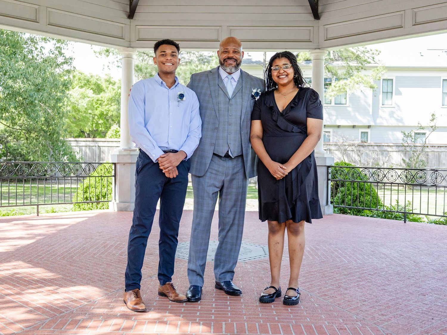Three people posing together under a gazebo on a brick patio, dressed in formal attire for a special occasion.