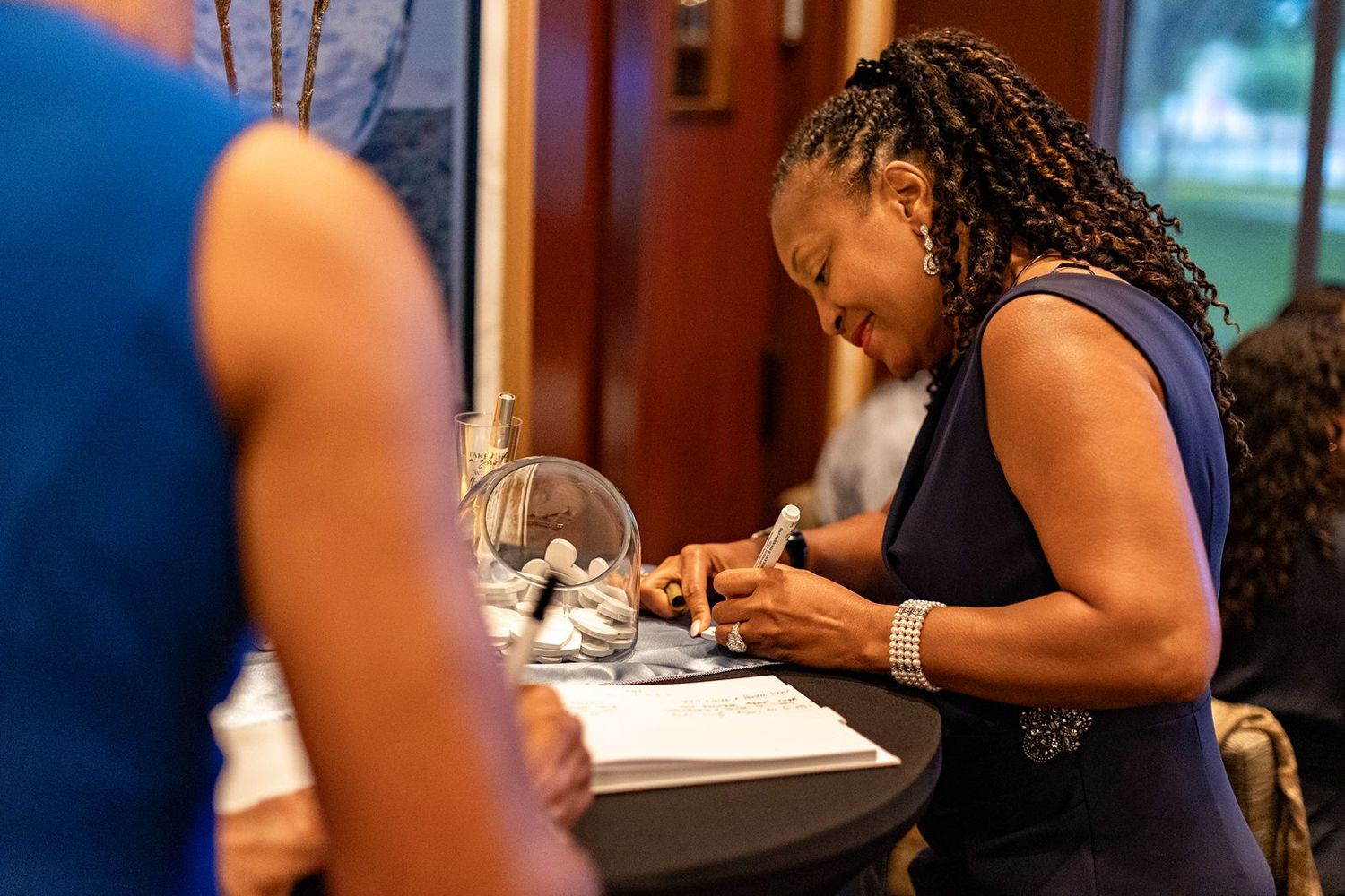 Woman in navy dress signs documents at a wedding guest book table during a reception.