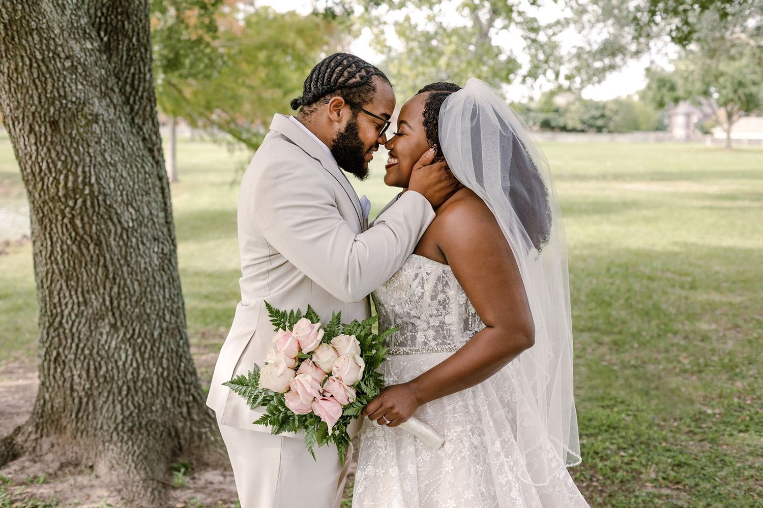A bride and groom share an intimate moment under a tree, with the bride holding a pink bouquet in a lace wedding gown.