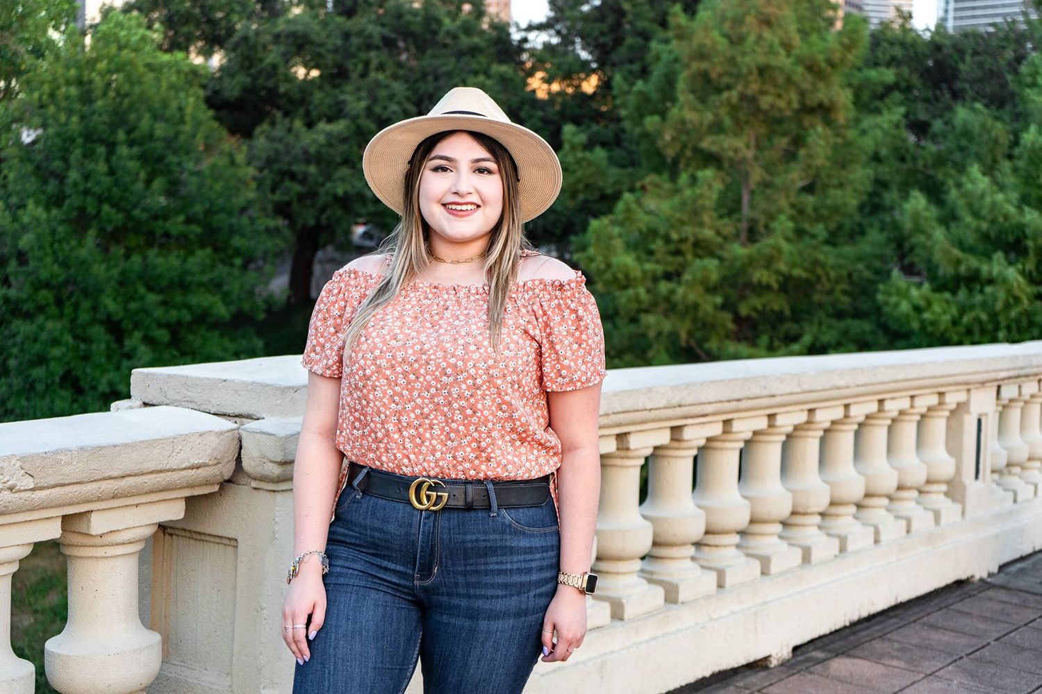 A woman in a peach polka dot top and straw hat poses on a stone balcony with lush greenery in the background.