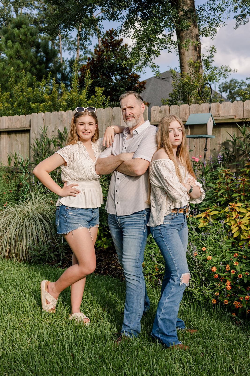 Three people in casual summer outfits pose playfully together in a backyard garden with yellow flowers and greenery.