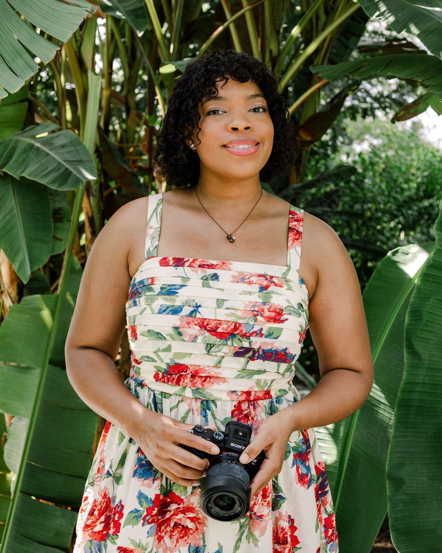 Woman in floral dress holding a camera, standing among lush tropical green leaves outdoors.