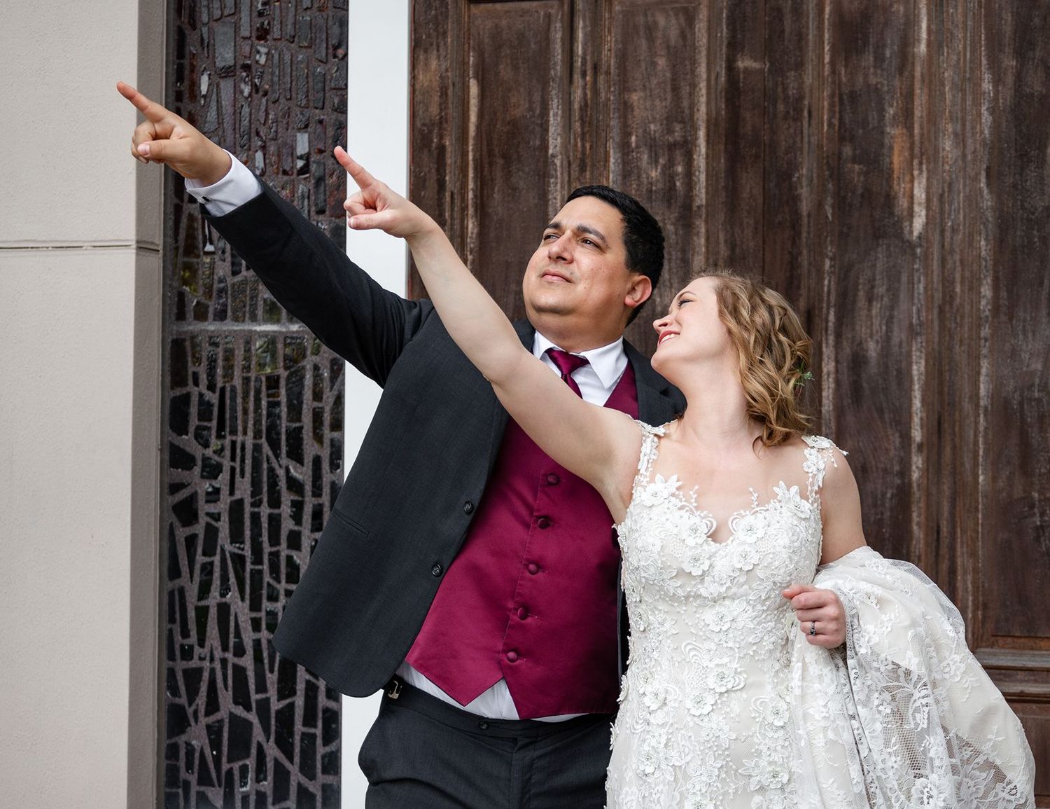 Bride and groom in wedding attire pointing and looking up joyfully outside a church door.