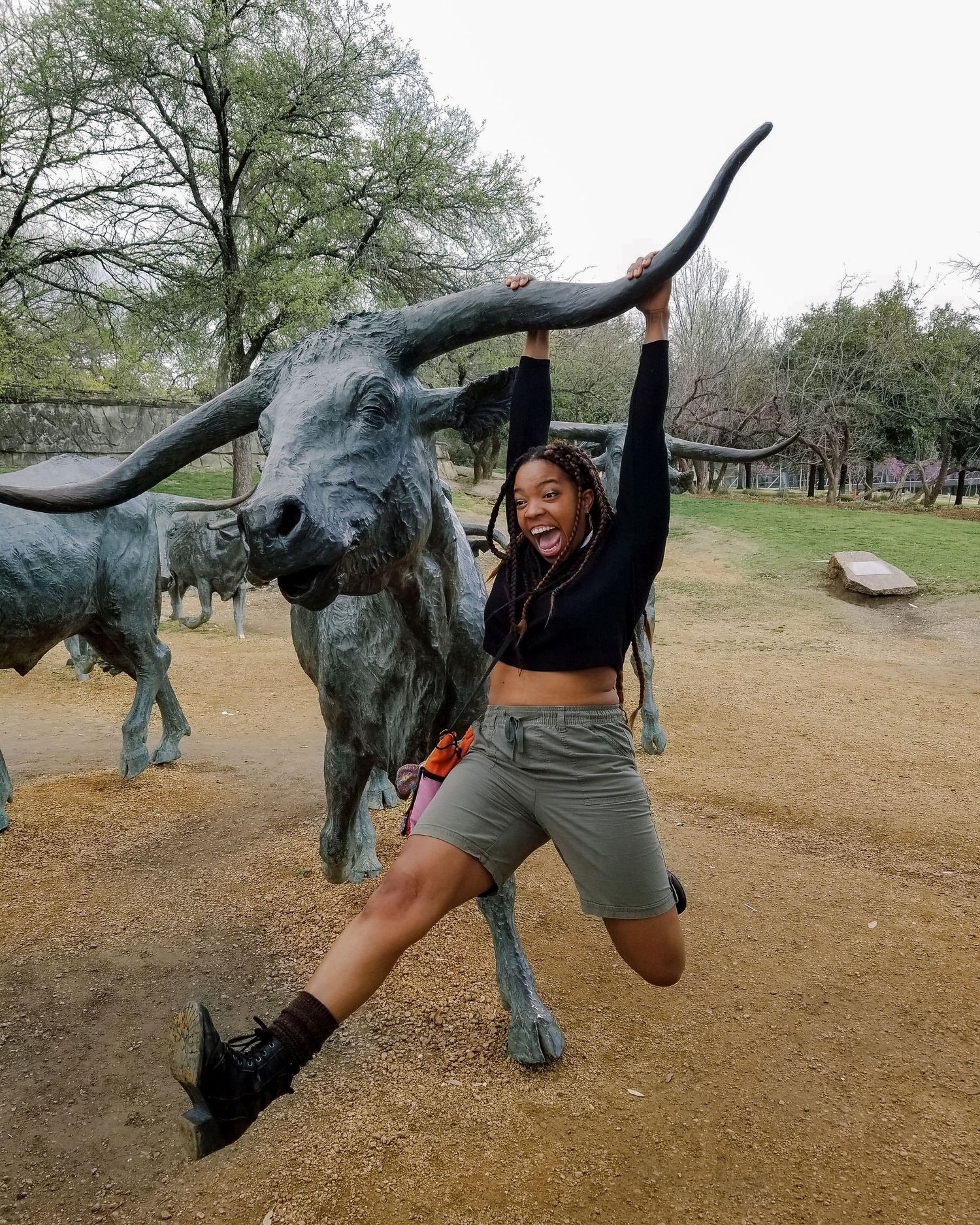 A joyful woman poses hanging from the horn of a bronze longhorn cattle sculpture outdoors in a park.