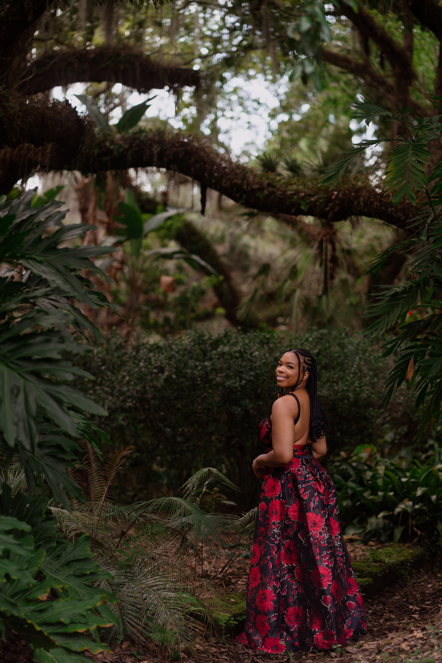 A woman in a red floral gown poses gracefully in a lush tropical garden with mossy trees.
