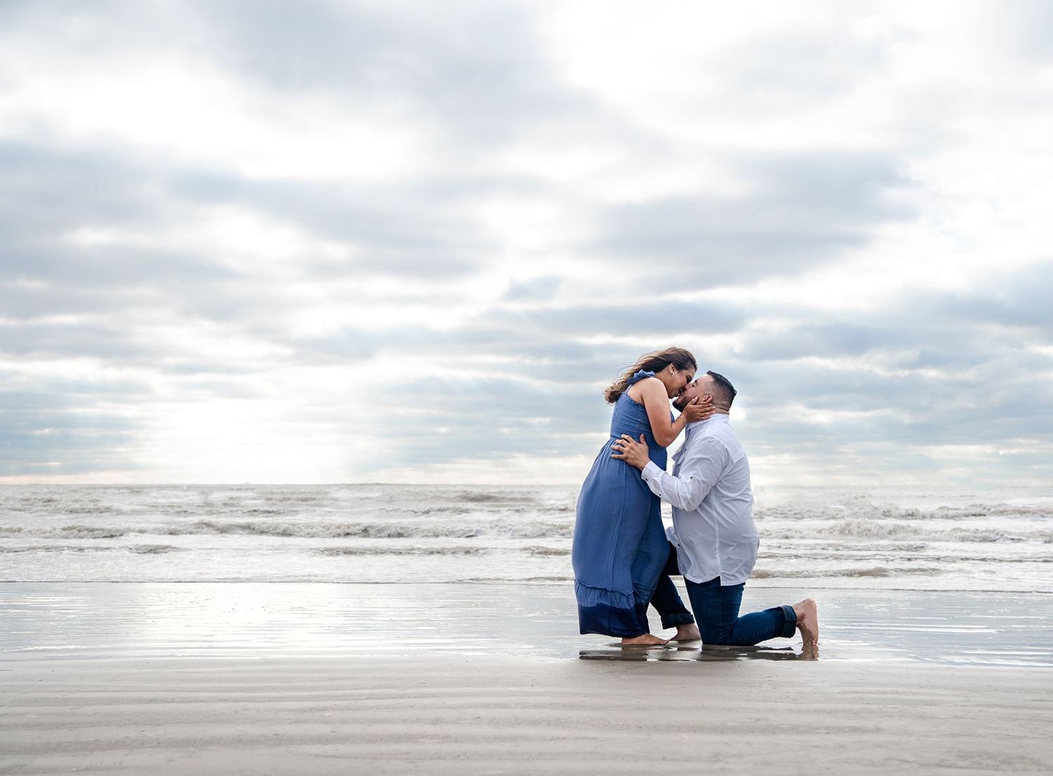 A romantic beachside moment between two people in blue and white outfits on a cloudy day by the ocean.
