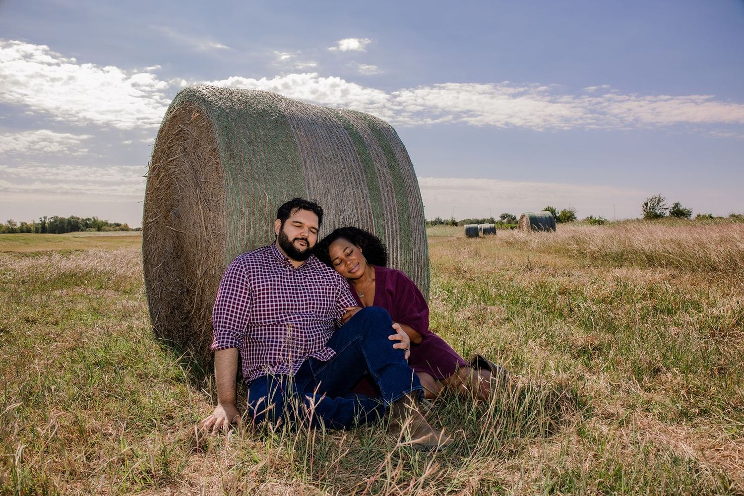 A couple in casual attire sits together against a large round hay bale in a golden rural field at sunset.