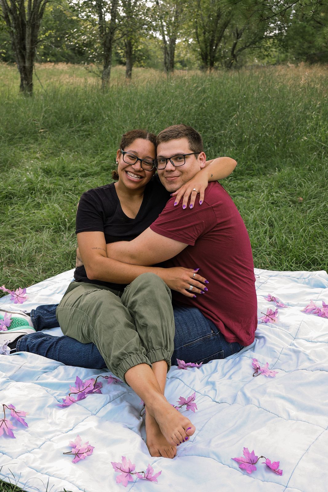 A smiling engaged couple embraces on a white blanket decorated with pink flowers in a green meadow.