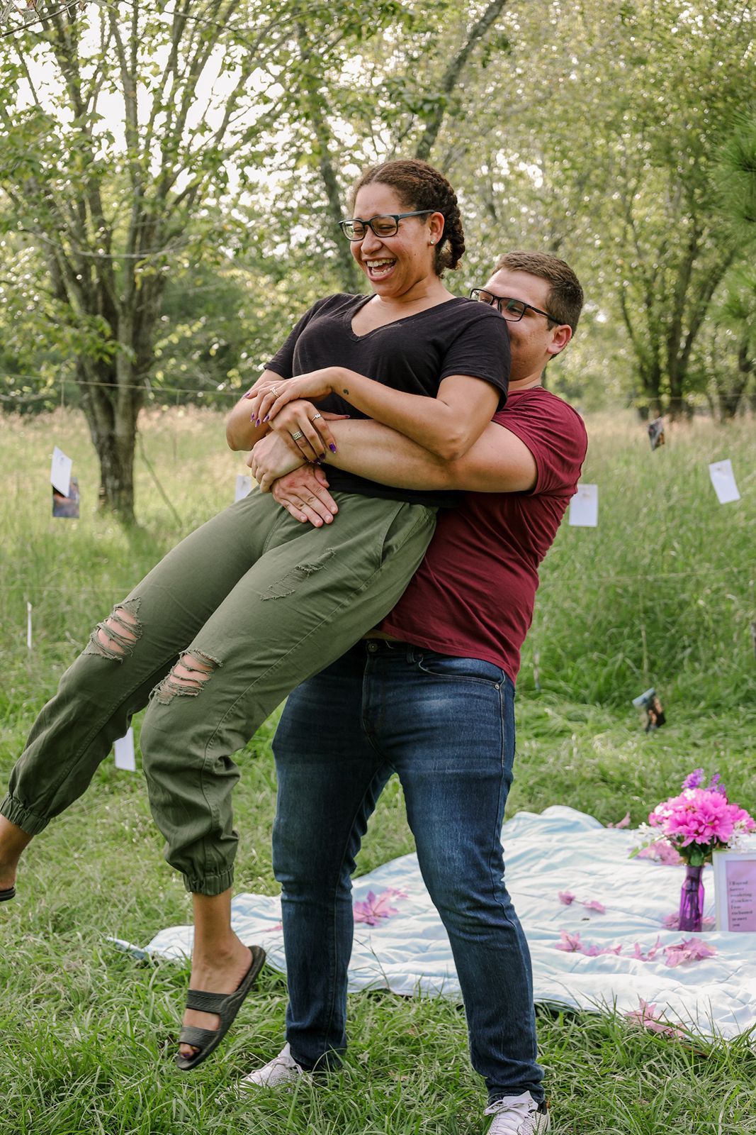 Couple laughing outdoors, man lifting woman in green pants near picnic blanket with pink flowers in park.