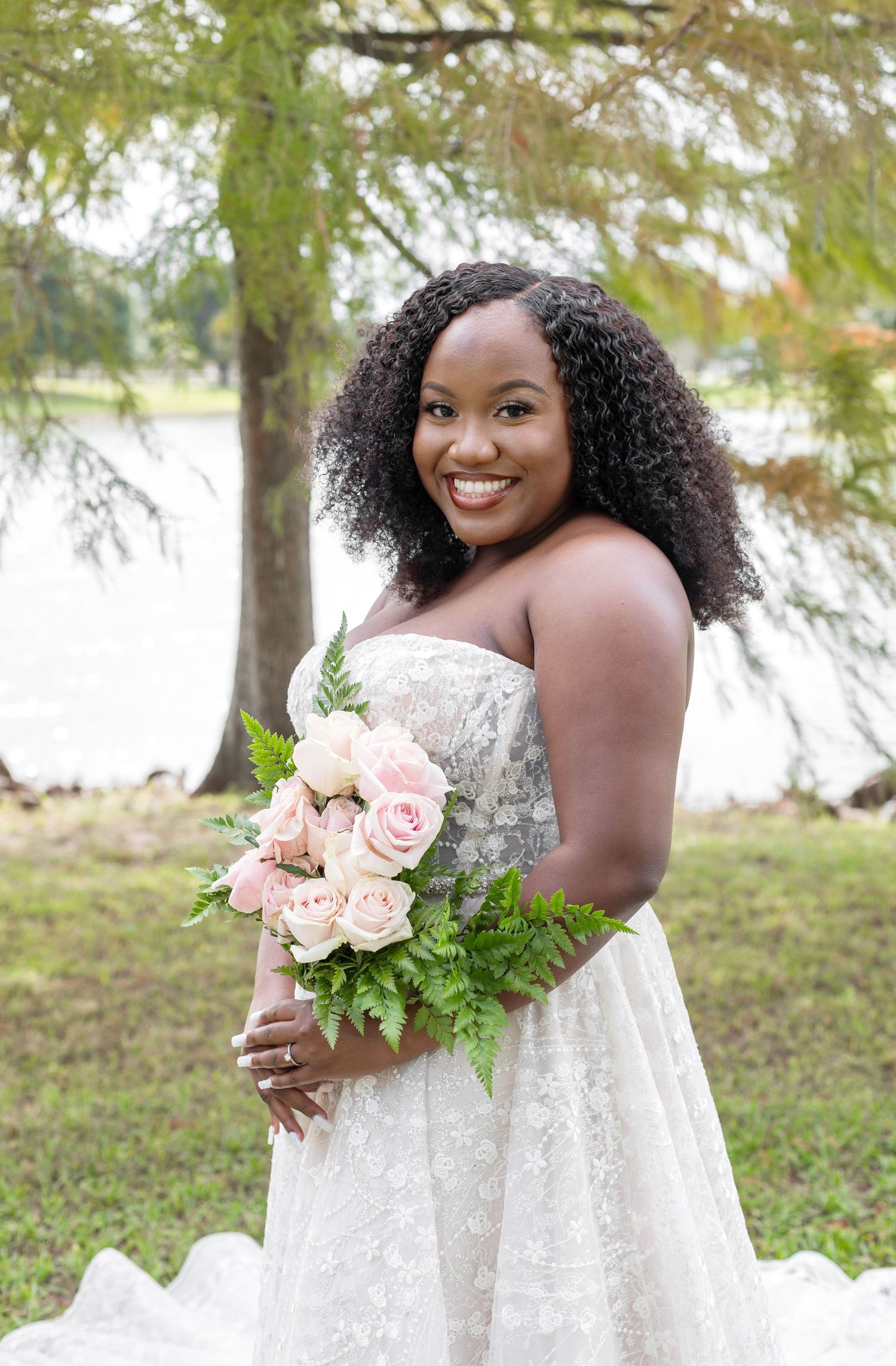 A bride in a white lace dress poses with a pink and green floral bouquet in an outdoor garden setting.