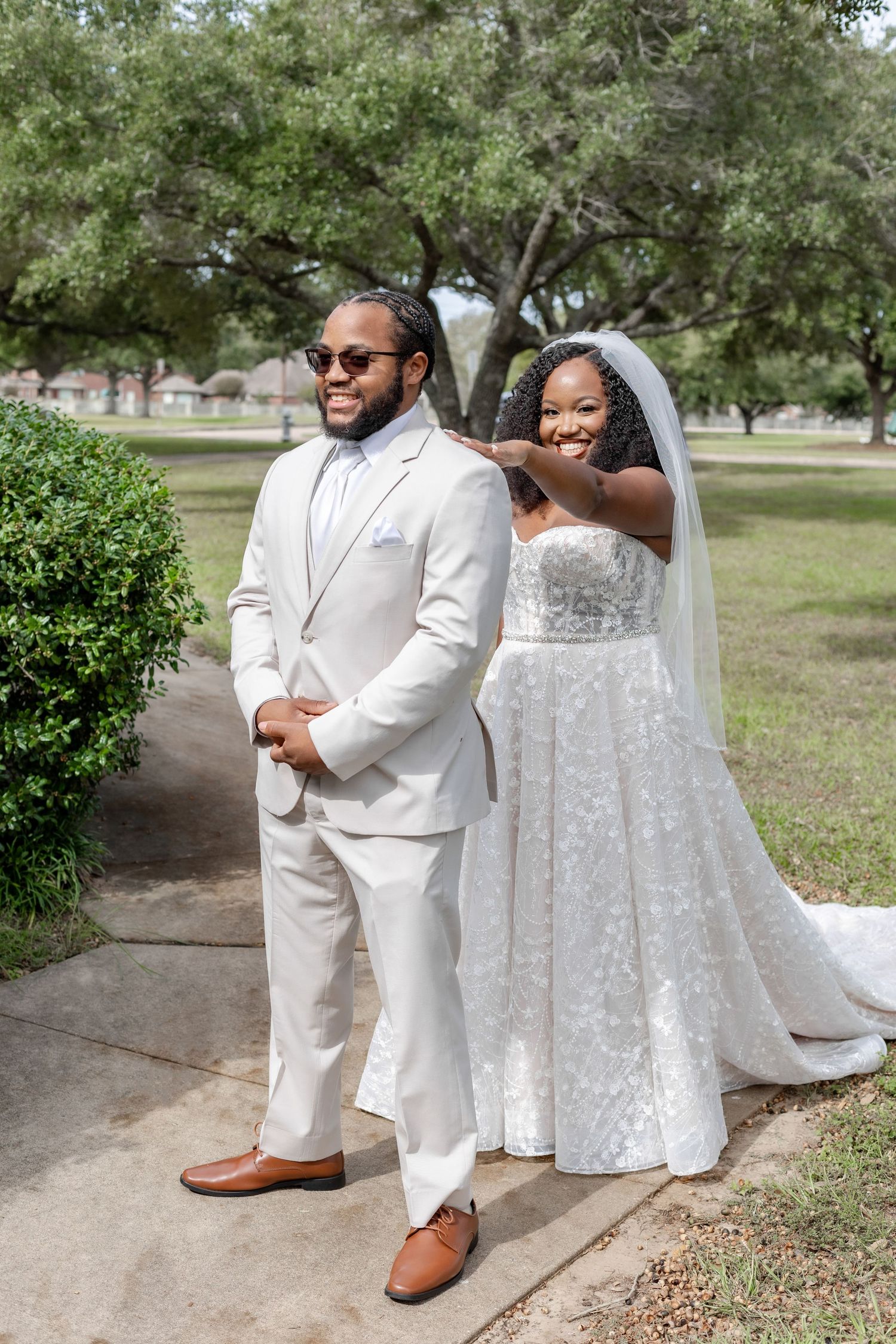 A wedding couple stands in front of lush greenery wearing elegant white attire and embracing.