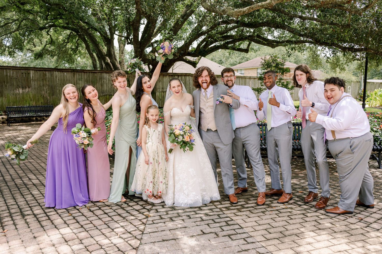 Wedding party in purple and grey attire posing outdoors under tree canopy.