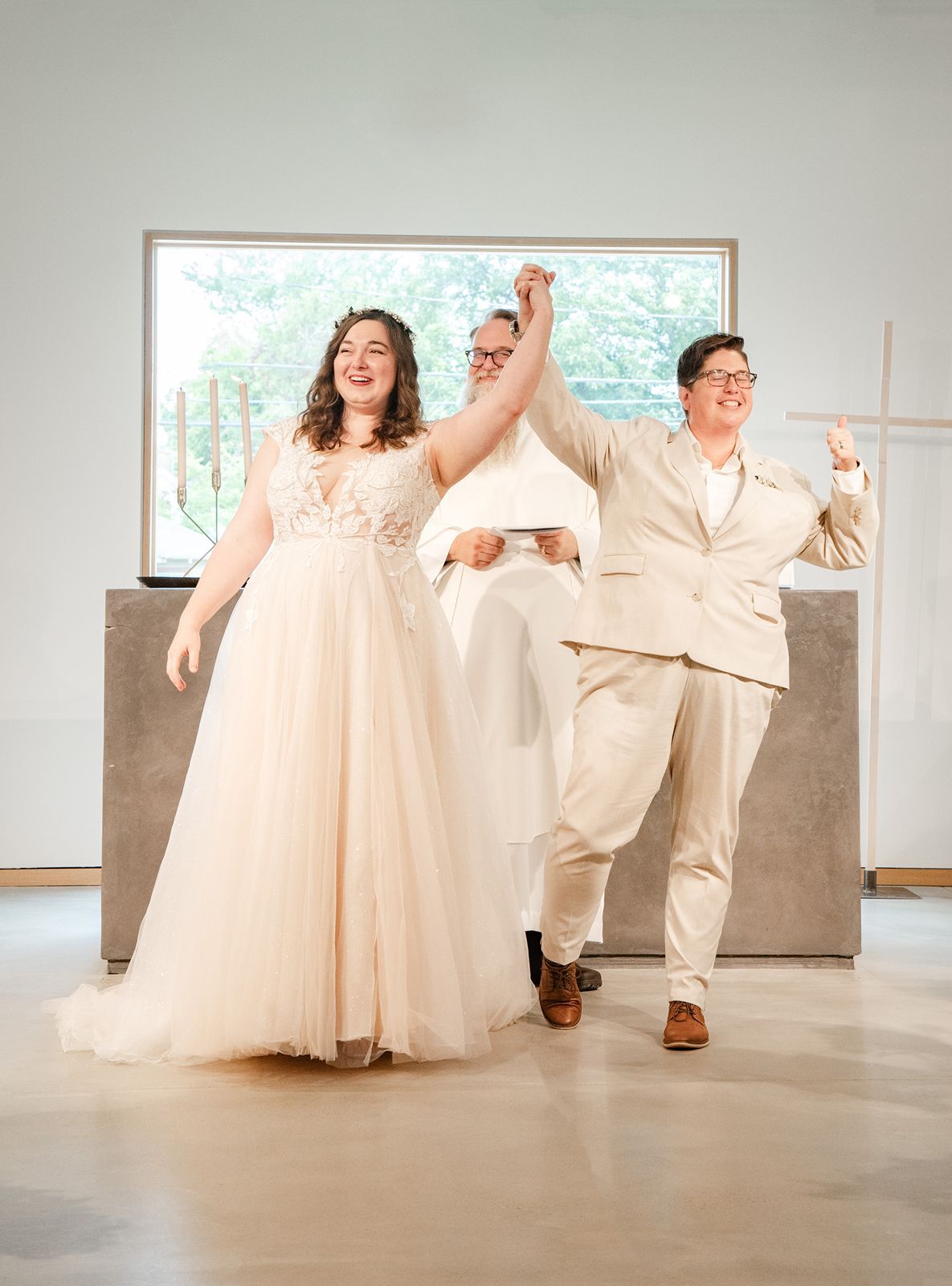 Two newlyweds in wedding attire celebrate joyfully with officiant raising their hands at a bright modern wedding venue.