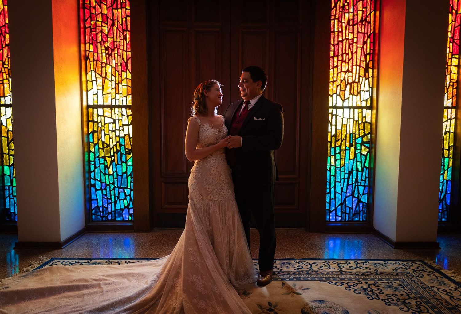 Bride and groom share a romantic moment surrounded by vibrant stained glass windows inside a church.