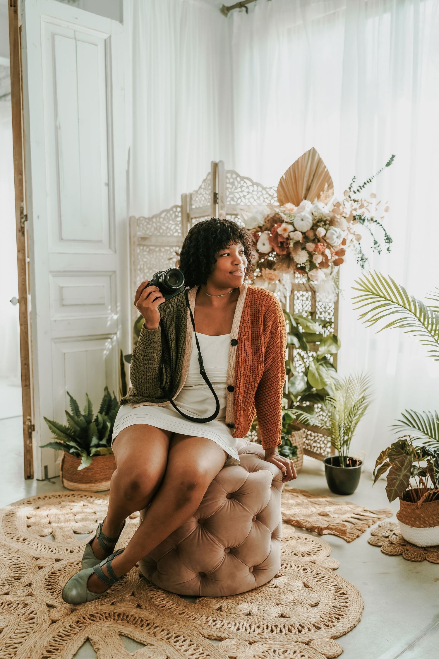 A young Black woman sits on a tufted ottoman holding a camera, wearing a white dress and brown cardigan, surrounded by tropical plants.