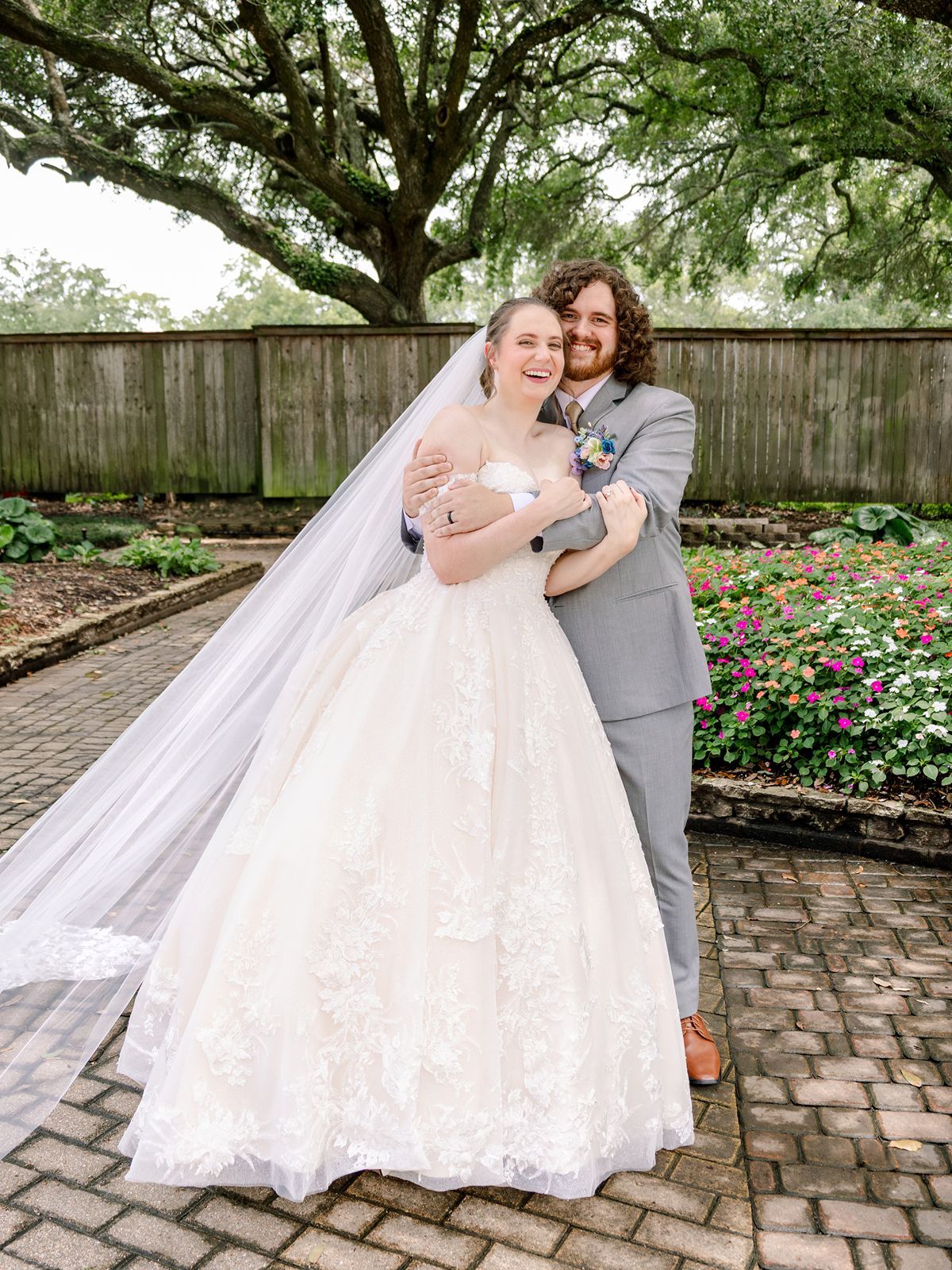 Smiling bride in lace ball gown with veil and groom in gray suit embracing in a garden courtyard.
