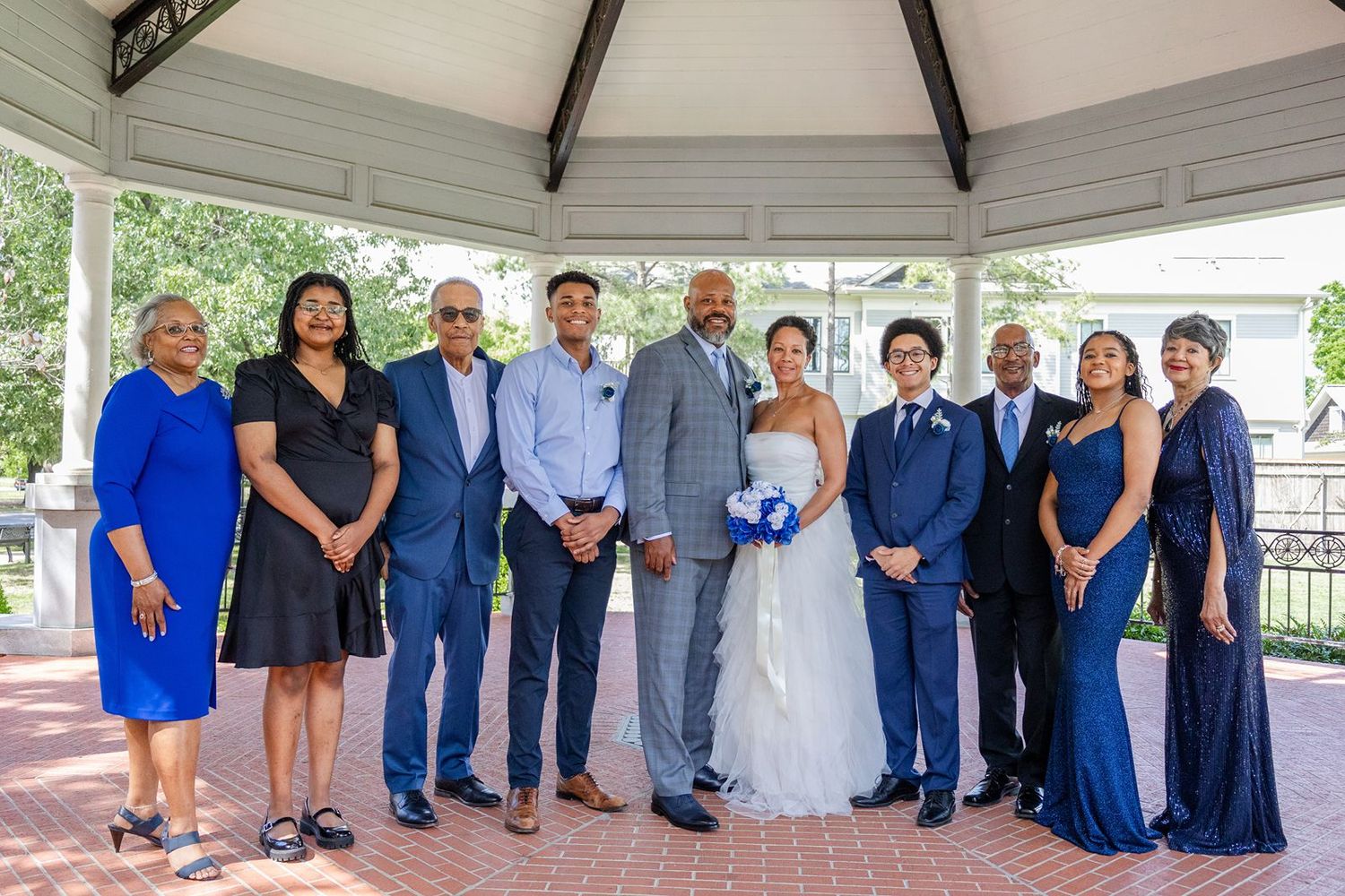 Wedding party group portrait with bride and groom surrounded by family in blue attire at gazebo.