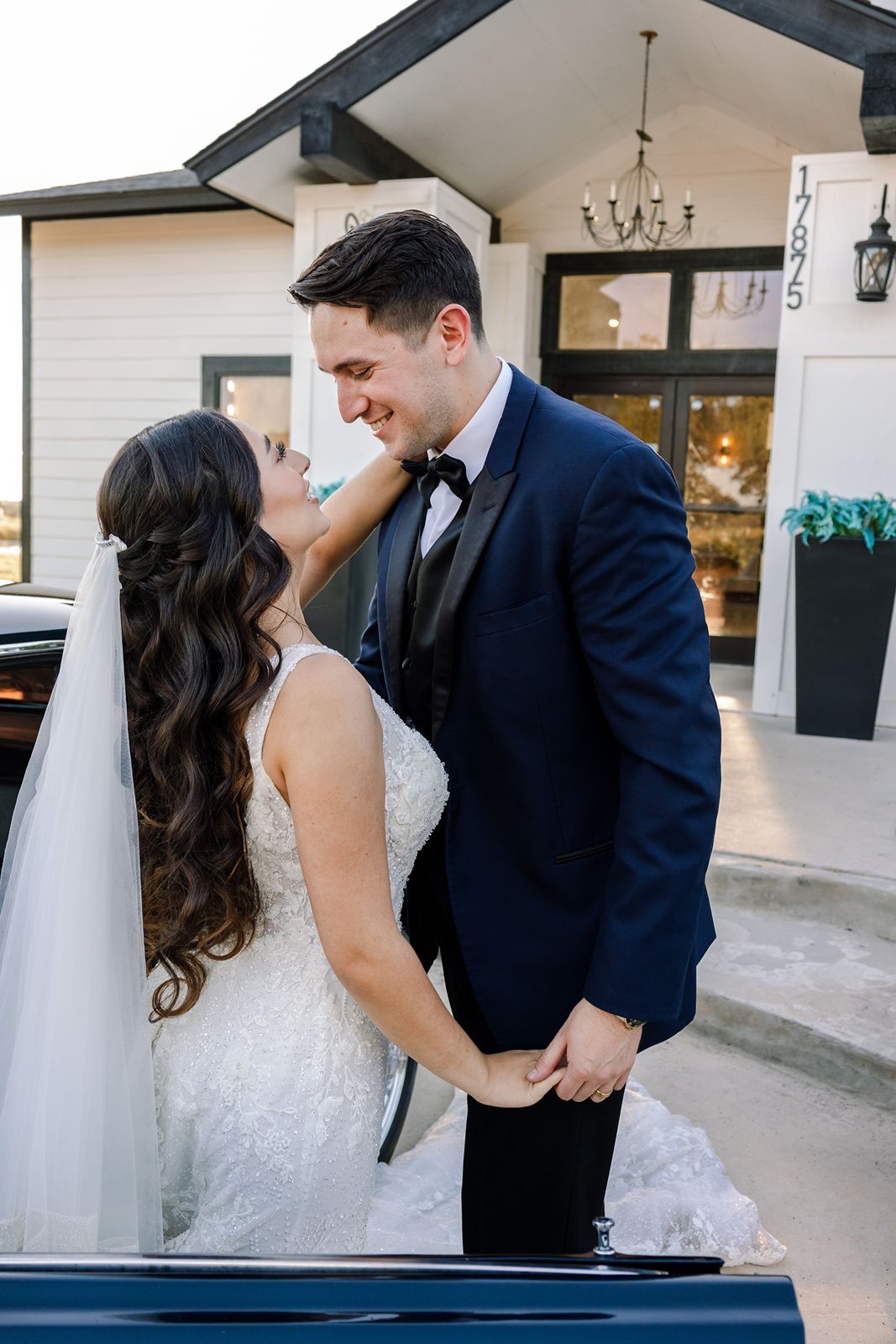 Bride and groom holding hands and smiling at each other outside a white wedding venue at dusk.