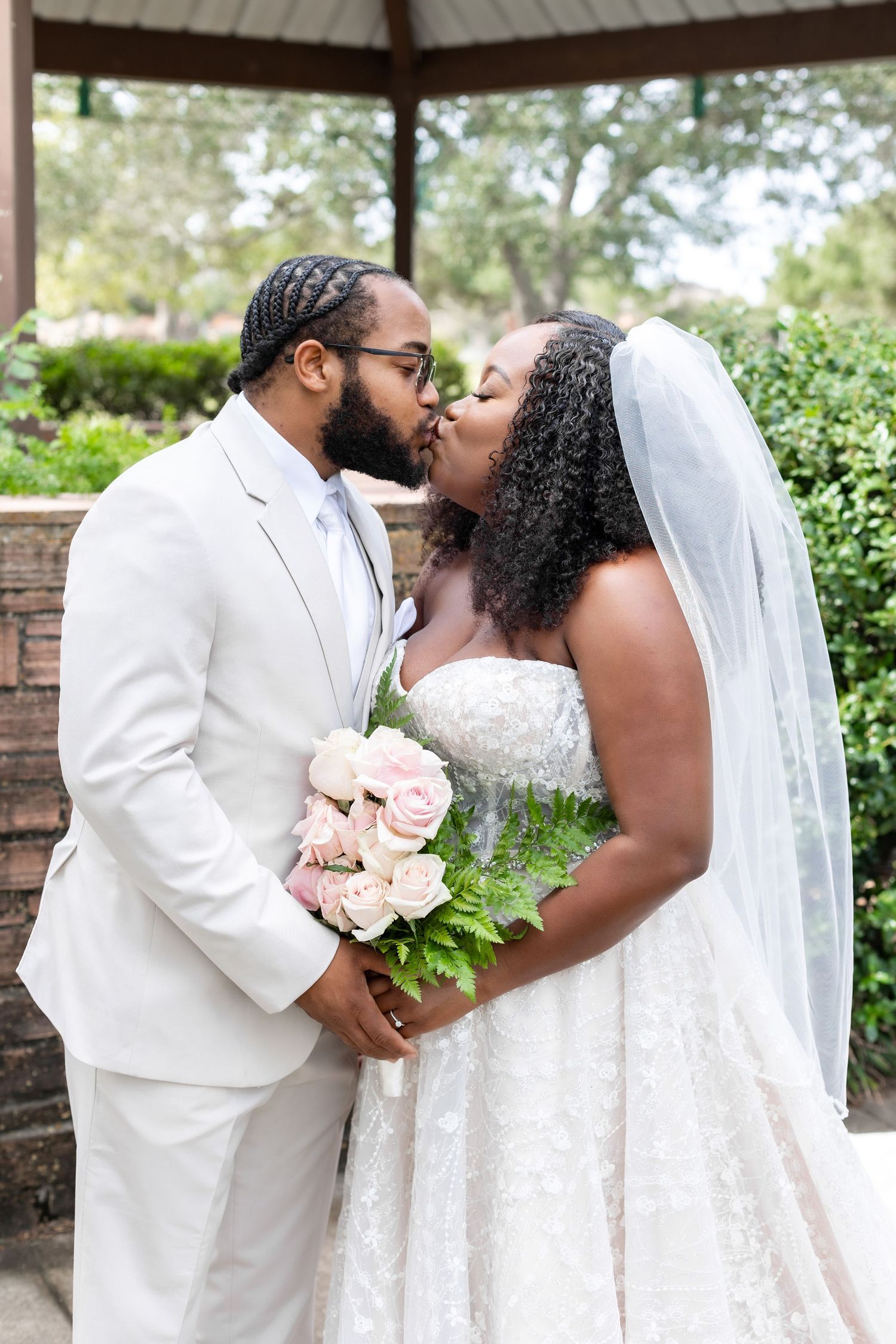 A tender wedding kiss moment under a gazebo with the bride holding a pink floral bouquet.