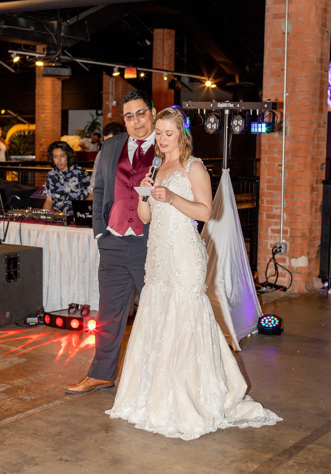 Bride and groom share a microphone during wedding reception speech in rustic brick venue with DJ setup behind them.