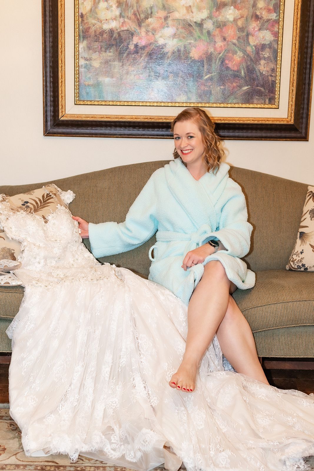 Smiling bride in light blue robe sits on sofa beside her lace wedding gown before getting dressed.